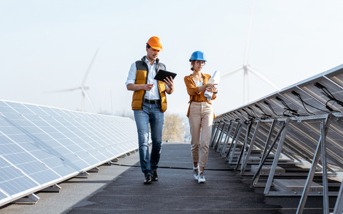 magazin-solarpark-kaufen-mobil Frau und Mann gehen zwischen zwei Reihen von aufgeständerten Solarmodulen entlang